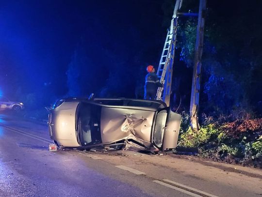 przewrócona toyota na leży na lewym boku, energetyk wchodzi po drabinie opartej o słup