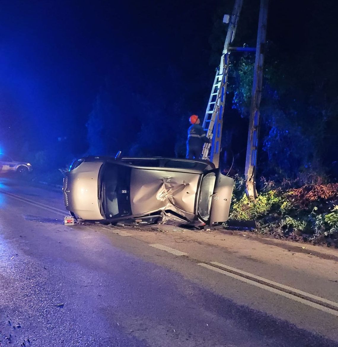 przewrócona toyota na leży na lewym boku, energetyk wchodzi po drabinie opartej o słup